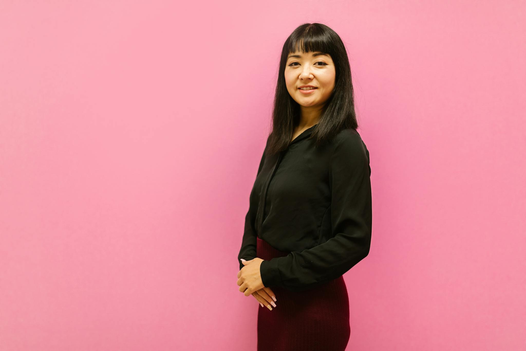 Asian woman in business attire smiling and standing against a pink background.