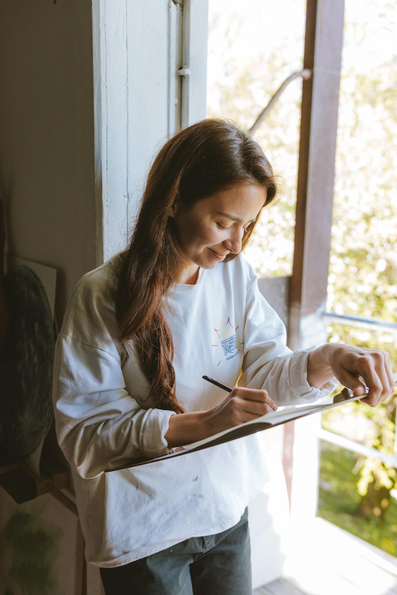 Smiling woman drawing on a clipboard by a sunlit window indoors.